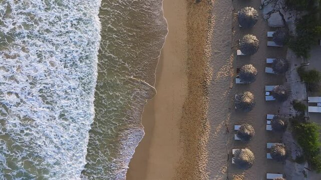Aerial view of a tranquil beach in Ayia Napa, Cyprus, with golden sand, gentle waves, thatched umbrellas, sun loungers, and a lone figure casting a shadow.