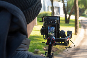 Vista lateral  trasera, de joven con cámara en estabilizador, filmando escena al aire libre, primer plano, enfoque selectivo, mano en máquina