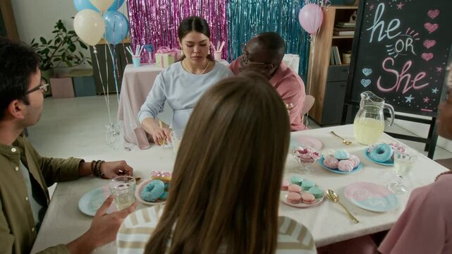 Medium arc shot of pregnant Caucasian woman enjoying dessert at gender reveal party, cheerful African American man stroking her belly, playfully teasing wife, and friends laughing