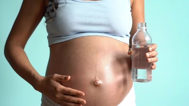 Pregnant Woman Hydrating with Water Bottle Gently Rubbing Belly in Pale Blue Top Promoting Maternal Health and Hydration Against a Soothing Background