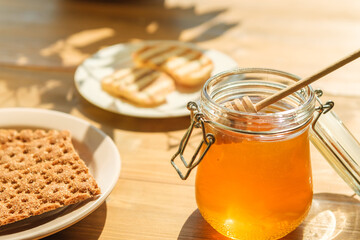 A glass jar filled with golden honey sits open beside a plate of freshly baked cookies on a warm wooden table, illuminated by afternoon sunlight.