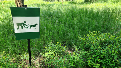 A green sign indicating dog walking rules in a grassy area. The sign features a person and a dog graphic, promoting cleaning up the excrement after the dog in the park, copy space