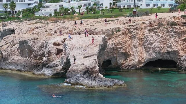 Rocky cliffs and turquoise waters in Ayia Napa, Cyprus, with visitors exploring, gentle waves, and resort buildings visible in the background.