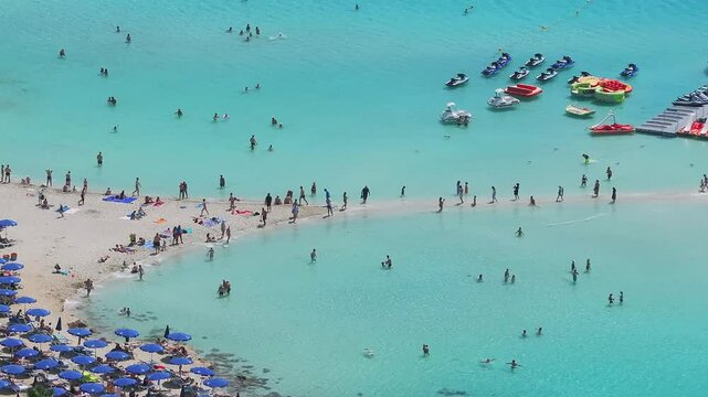 Turquoise waters with swimmers, jet skis, and inflatables near a dock. Sandy Nissi beach lined with blue umbrellas and sun loungers, with people walking and relaxing.
