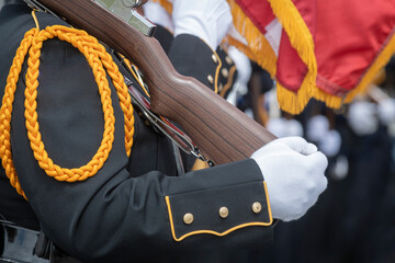 A uniformed person holds a rifle, wearing black attire with gold accents and a braided cord. 