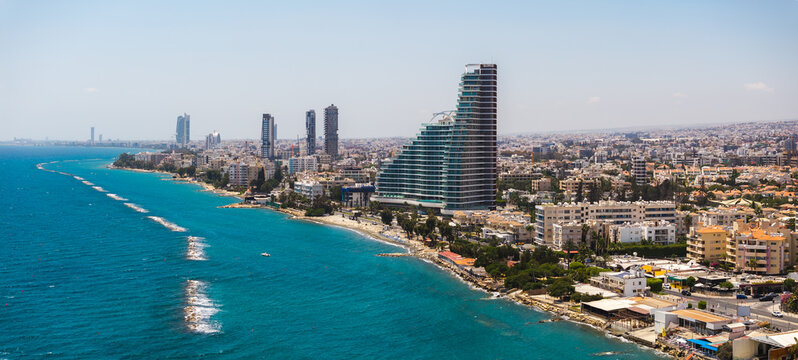 Aerial view of Limassol, Cyprus, featuring modern high rise buildings, a curved structure, turquoise sea, sandy beach, and wave breakers.