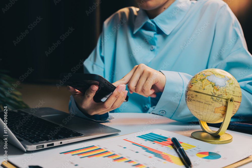 Poster person using smartphone while working on laptop with charts, globe, and data analysis on table in mo