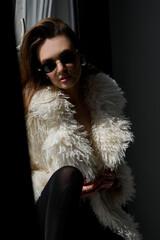 Beautiful girl in a fur coat and sunglasses sits on the windowsill