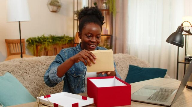 African American young woman sitting at table on home sofa happily checks envelope full of cash dollars received as a birthday gift. Black girl opens big gift box and smiles with joy and surprise.