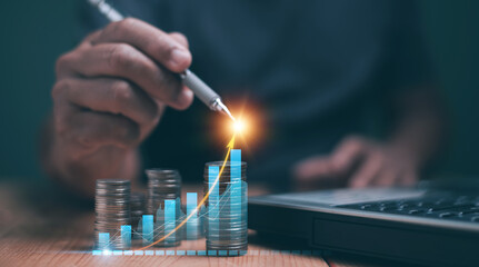 Business analytics and financial concept. Saving money for future growth. Businessman using pen pointing to analyze financial graph for investment, Stack of coins and laptop. Financial planning.