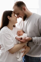 Portrait of a young family with infant in his arms photographed on a light window background.