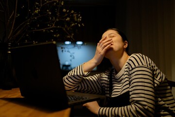 A woman sitting at a table with a laptop, yawning. She is wearing a striped shirt and has her hair...