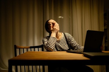 A woman sitting at a table with a laptop, yawning. She is wearing a striped shirt and has her hair...