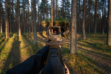 Aiming an assault rifle with a holographic sight in a forest, first-person view, tactical training...
