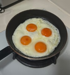 Four sunny side up eggs cooking in a black frying pan on a stove. Bright orange yolks and perfectly set whites in a simple kitchen setting