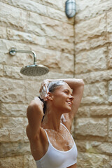 woman showering outdoors with wet hair, smiling and relaxed under rain showerhead against stone...