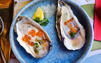 Top viiew of Tasmanian Oyster topped with salmon eggs, sliced spring onions, and lemon on a blue dish. Seafood on plate.