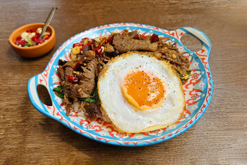 Stir fried beef with basil and dried chili paste over white rice with Thai fried egg on wooden background. Flat lay of Spicy and famous food in Thailand with fish sauce in small bowl on brown table.