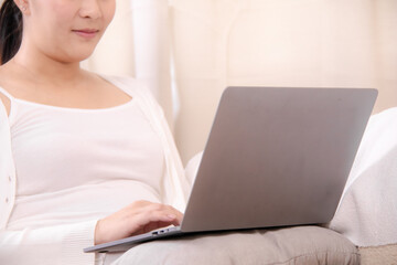 Fototapeta premium Pregnancy working, pregnant Asian woman using a laptop at home while gently touching her belly, combining productivity and care, small business owner, work-from-home, or maternity service promotions