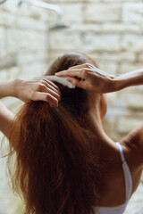 Naklejka premium Woman brushing hair with white comb, natural light, brown long hair, casual white strap top, textured stone wall background in cozy indoor setting.