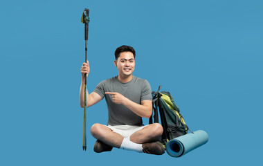 Enthusiastic hiker with a backpack and trekking pole seated and pointing on a solid backdrop