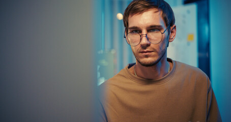 Obraz premium Caucasian male engineer looks at computer screen in modern tech office at night. Deep concentration during software analysis and programming task.