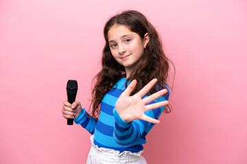 Little singer girl picking up a microphone isolated on pink background saluting with hand with...