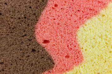 Close up captures the textured surface of a three layered sponge cake. The distinct colors a rich brown, a warm pink, and a bright yellow merge with soft, undulating lines.