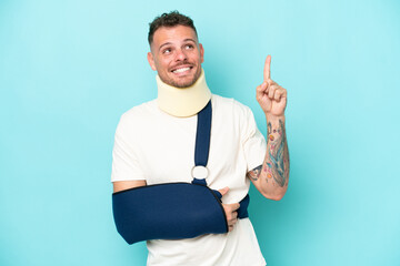Young caucasian man wearing a sling  and neck lace isolated on blue background pointing up a great...