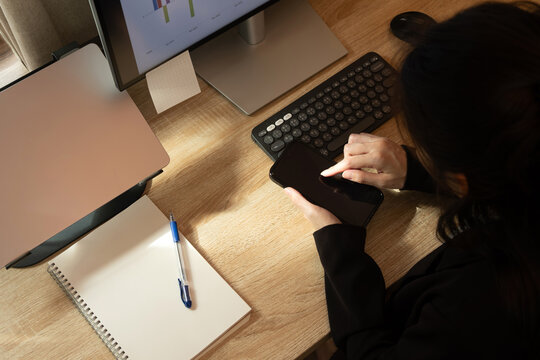 Woman Focused on Smartphone at Office Desk with Laptop and Data Monitor.