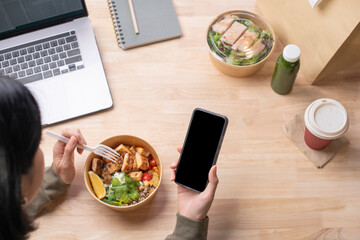 Asian woman eating healthy meal while working, Businesswoman having a lunch break at the office, hand holding phone with blank black screen for copyspace