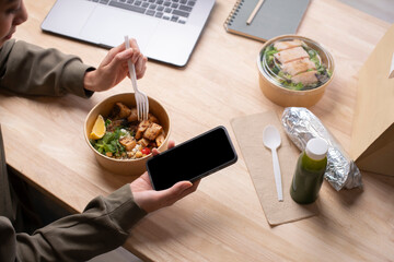 Asian woman eating healthy meal while working, Businesswoman having a lunch break at the office, hand holding phone with blank black screen for copyspace