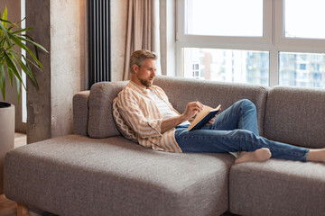 Young businessman sitting on sofa at home and writing notes down on a notepad