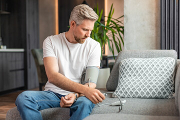 Fototapeta premium Fair-haired, bearded guy checking blood pressure at home, using tonometer while sitting on couch in living room, copy space.