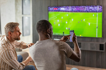 Two friends holding smartphone with sports bets app while watching football match at home in living room