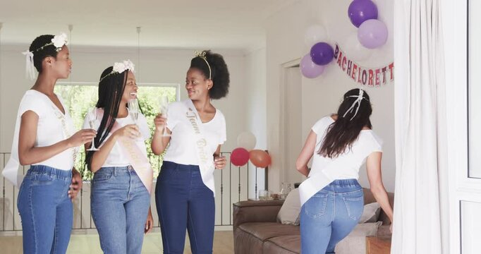 Diverse female friends at home wearing sashes clinking flutes celebrating bride-to-be entering