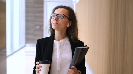 Confident businesswoman navigating modern office corridor, carrying coffee and documents, wearing wireless earbuds, embodying professional workplace dynamics and leadership presence