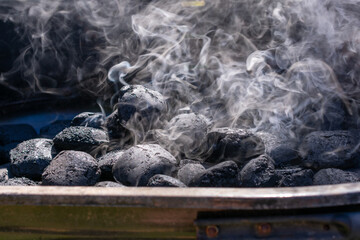 Close-up of charcoal briquettes heating in a grill with white smoke rising. The coals are still mostly black, indicating the early stage of lighting.
