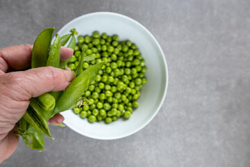 hand holding empty pea pods with white bowl of green peas on gray surface