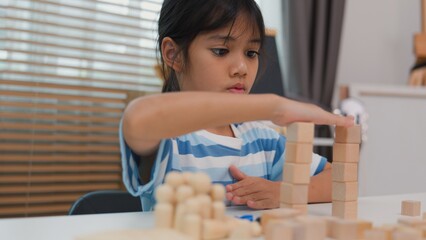 Young Asian girl focused on stacking wooden blocks, promoting STEM learning, creativity, and motor skill development. Concept of early education, problem solving, and cognitive growth.