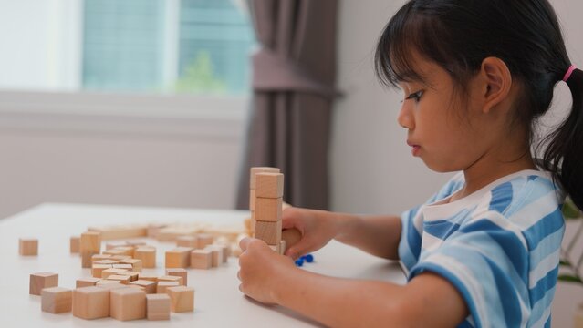 Young Asian girl focused on stacking wooden blocks, promoting STEM learning, creativity, and motor skill development. Concept of early education, problem solving, and cognitive growth.