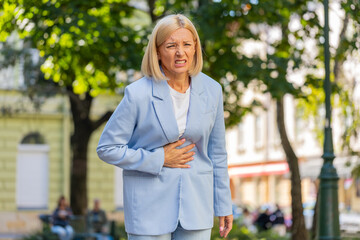 Sad mature businesswoman boss in formal suit suffering from stomach pain while walking on city town...