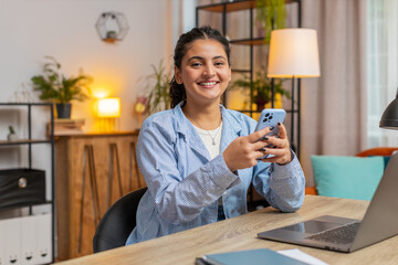 Happy young Indian woman sitting at table using smartphone smiling at home apartment. Girl...