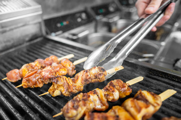 Close-up of skewered chicken grilling on a barbecue. A hand turns the skewers with tongs in a professional kitchen setting. Juicy, golden-brown pieces sizzling over open flames.