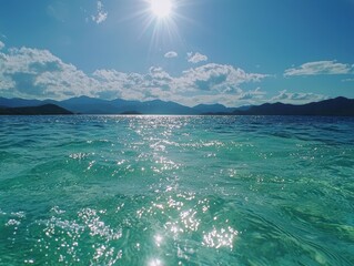 Clear turquoise ocean water sparkles under a bright sun with distant mountains and a blue sky filled with scattered clouds.