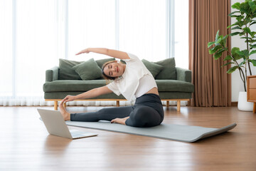 Young Asian woman stretching yoga workout on exercise mat while online training class with computer laptop in living room