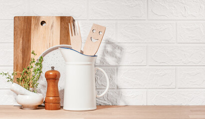 Kitchen utensils including a smiling wooden spatula, pepper mill, mortar with thyme, and cutting board arranged on a wooden countertop against a white brick wall for a cozy home setting