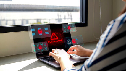 A person in a black shirt is typing on a keyboard with a futuristic transparent screen displaying red warning symbols, indicating a cybersecurity threat or hacking attempt