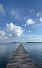 Obraz premium A long wooden jetty stretches straight out into the calm ocean, leading towards distant islands under a blue sky with clouds.