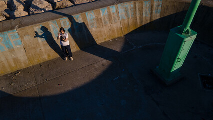 Young woman with a camera standing on a pier in late afternoon light. Creative urban composition with long shadows, concrete textures, and a graphic green structure.